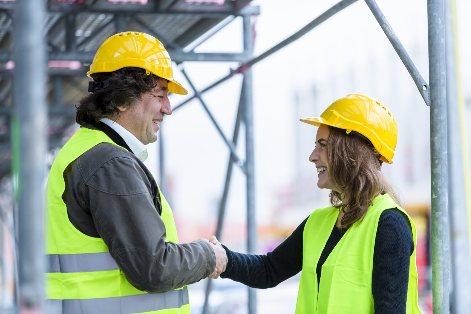 Two engineers, a man and a woman, with safety yellow jackets and hardhats, shaking hands